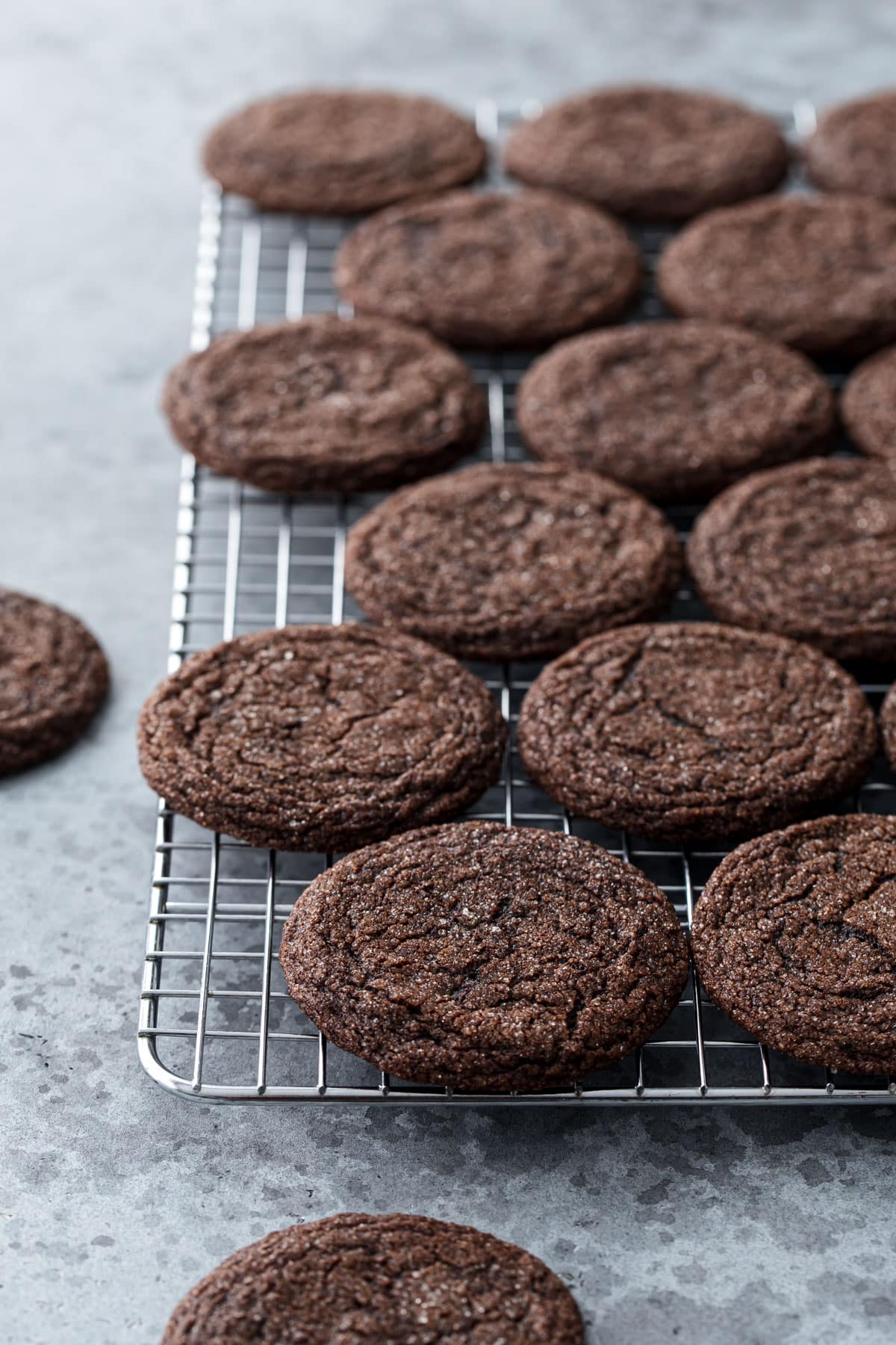 Chewy Chocolate Molasses Cookies on a wire baking rack, two cookies off to the side on a gray background.