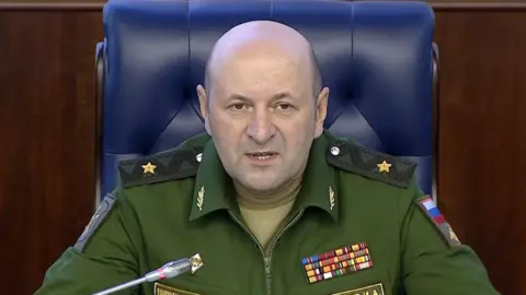 AP Igor Kirillov, who has a bald head and wears green military overalls, speaks into a mic during a press conference