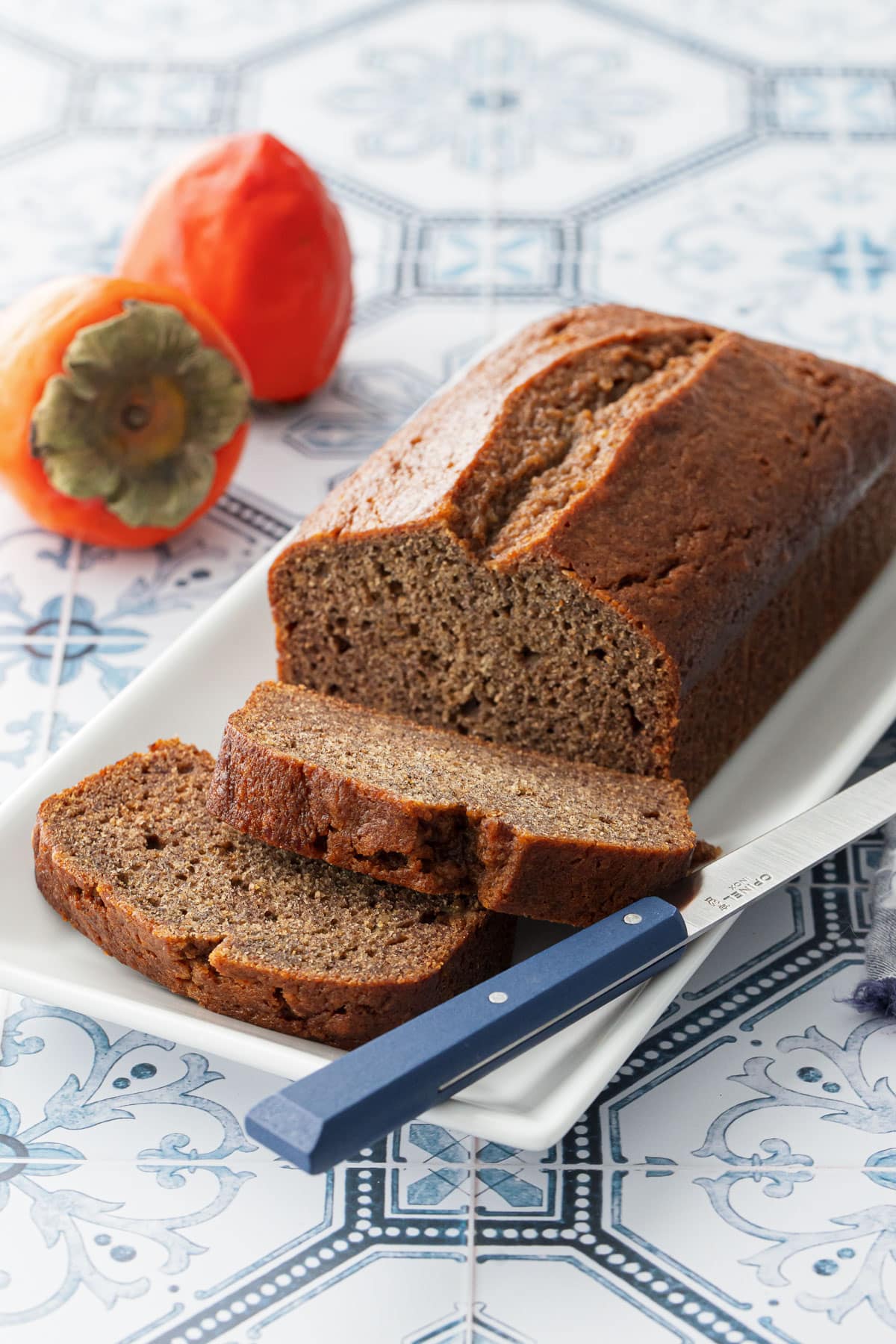 Loaf of Spiced Persimmon Bread on a white cake plate, two slices cut and laying down, with two hachiya persimmons in the background.