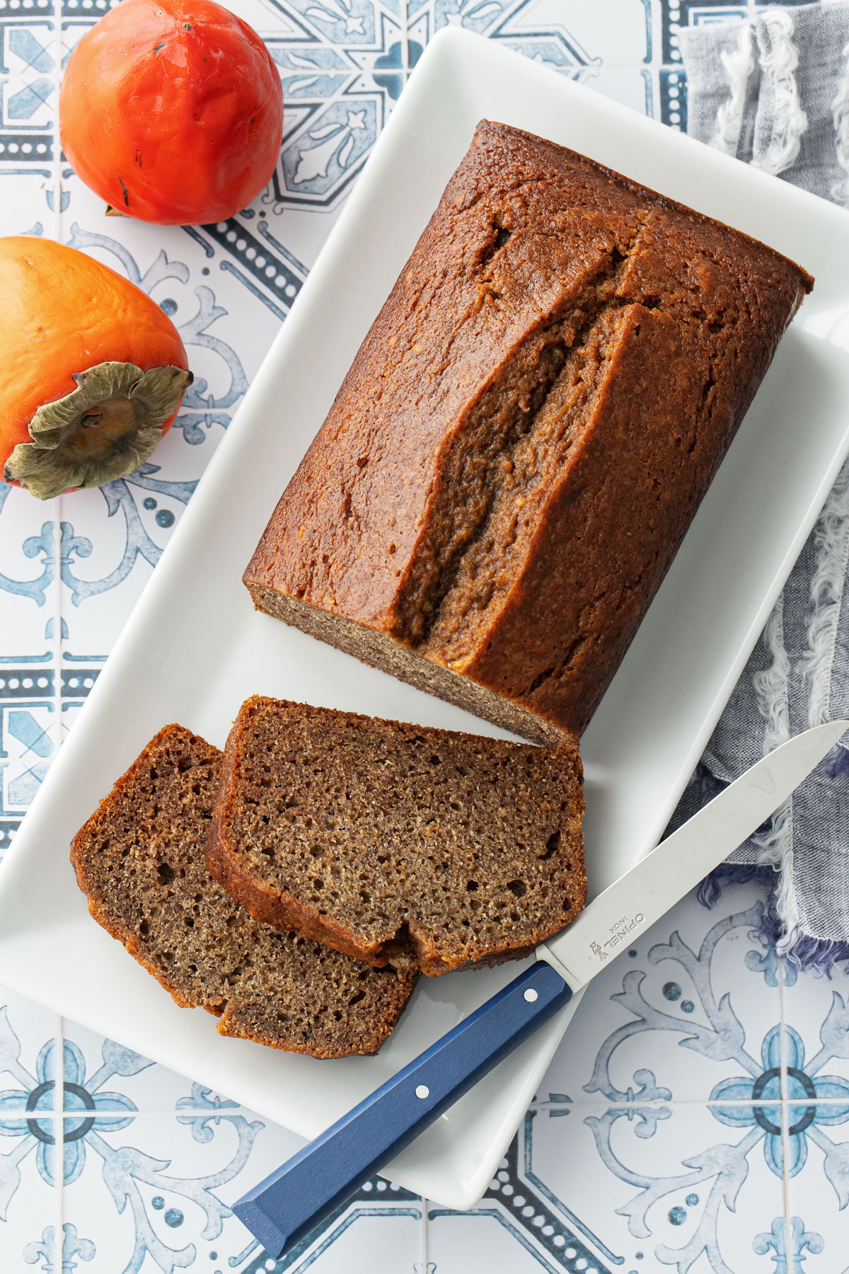 Overhead, Spiced Persimmon Bread loaf on a white plate with two slices cut and laying down, on a blue and white tile background with two orange persimmons on the side.