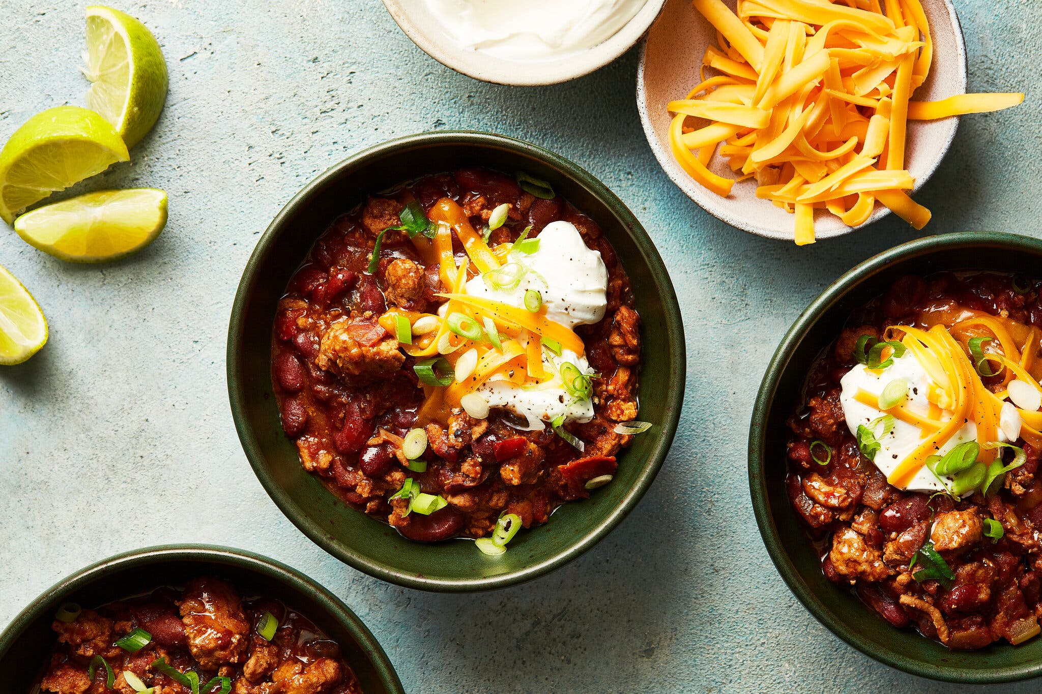 Three green bowls of turkey chili are topped with sour cream, shredded Cheddar and sliced scallions. Lime wedges and smaller bowls of additional cheese and sour cream are nearby.