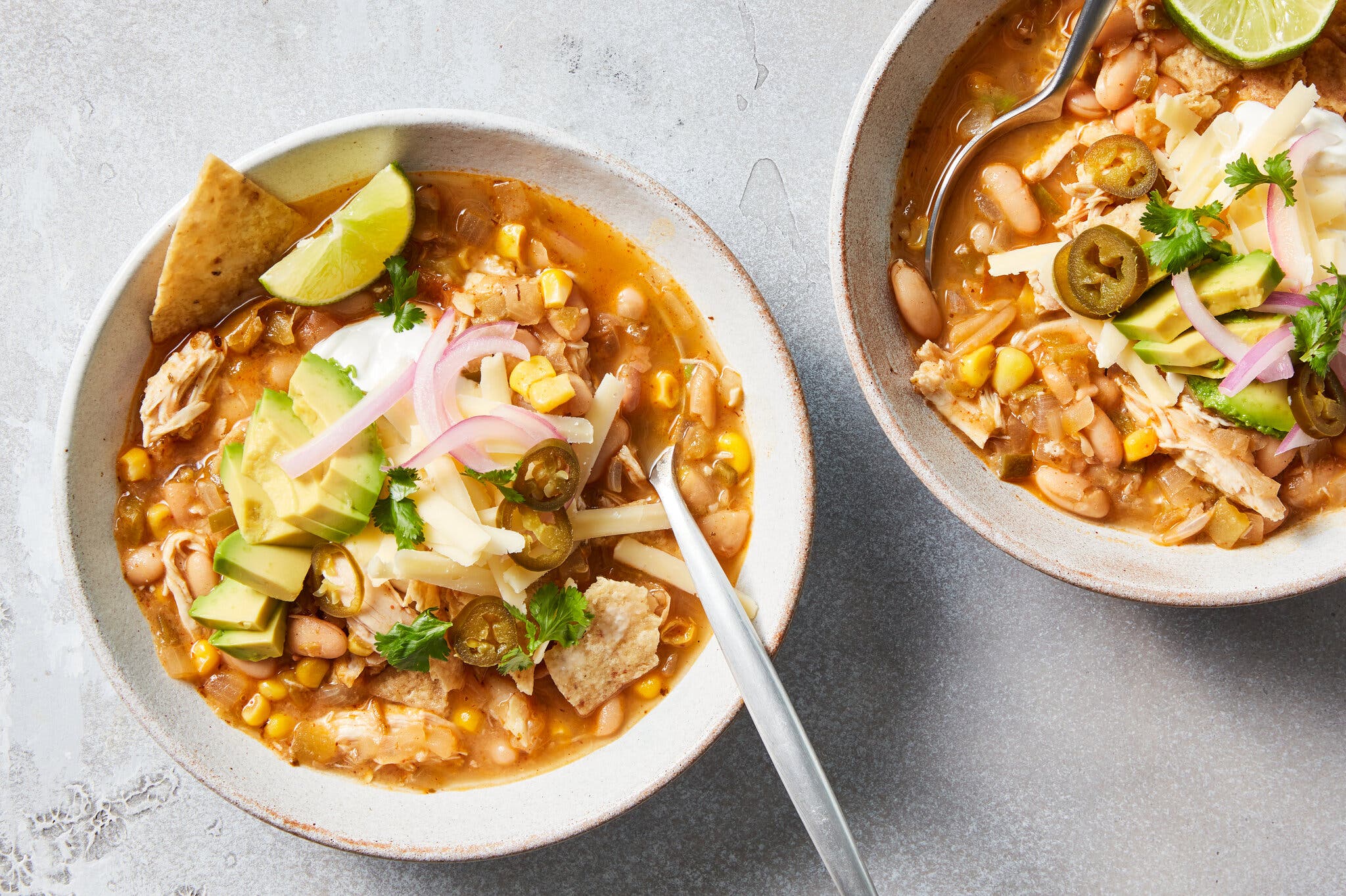 Two servings of white chicken chili are served in white bowls and topped with shredded cheese, slivered red onion, avocado slices, crushed tortilla chips and limes for squeezing.