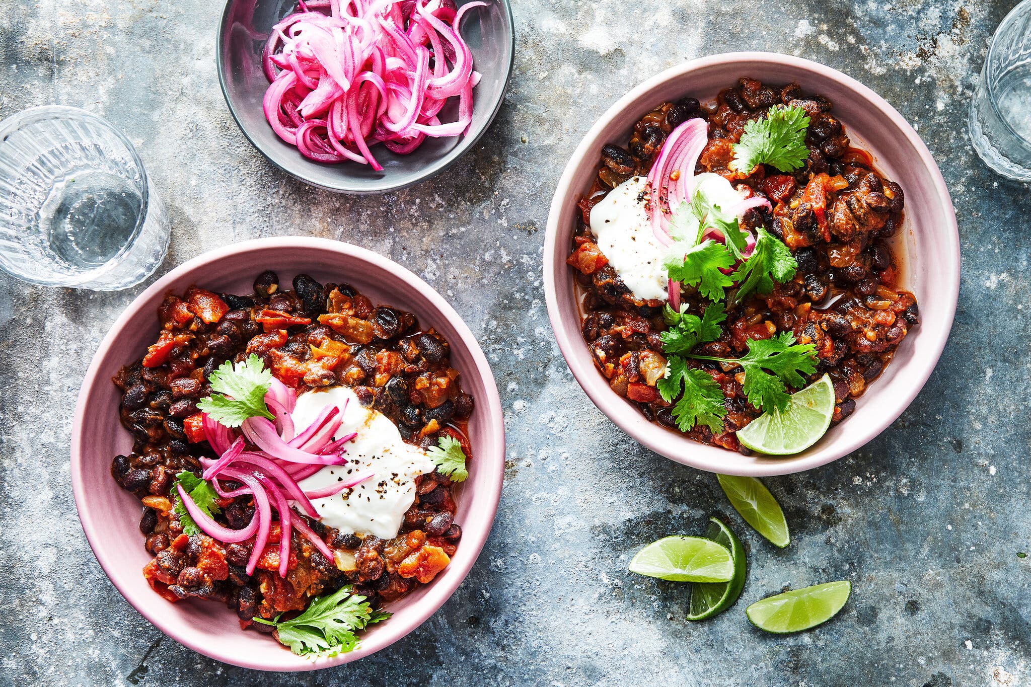 Two bowls of chili topped with sour cream, pickled red onions and herbs are photographed from overhead. A plate with more pickled onions sits off to the side.