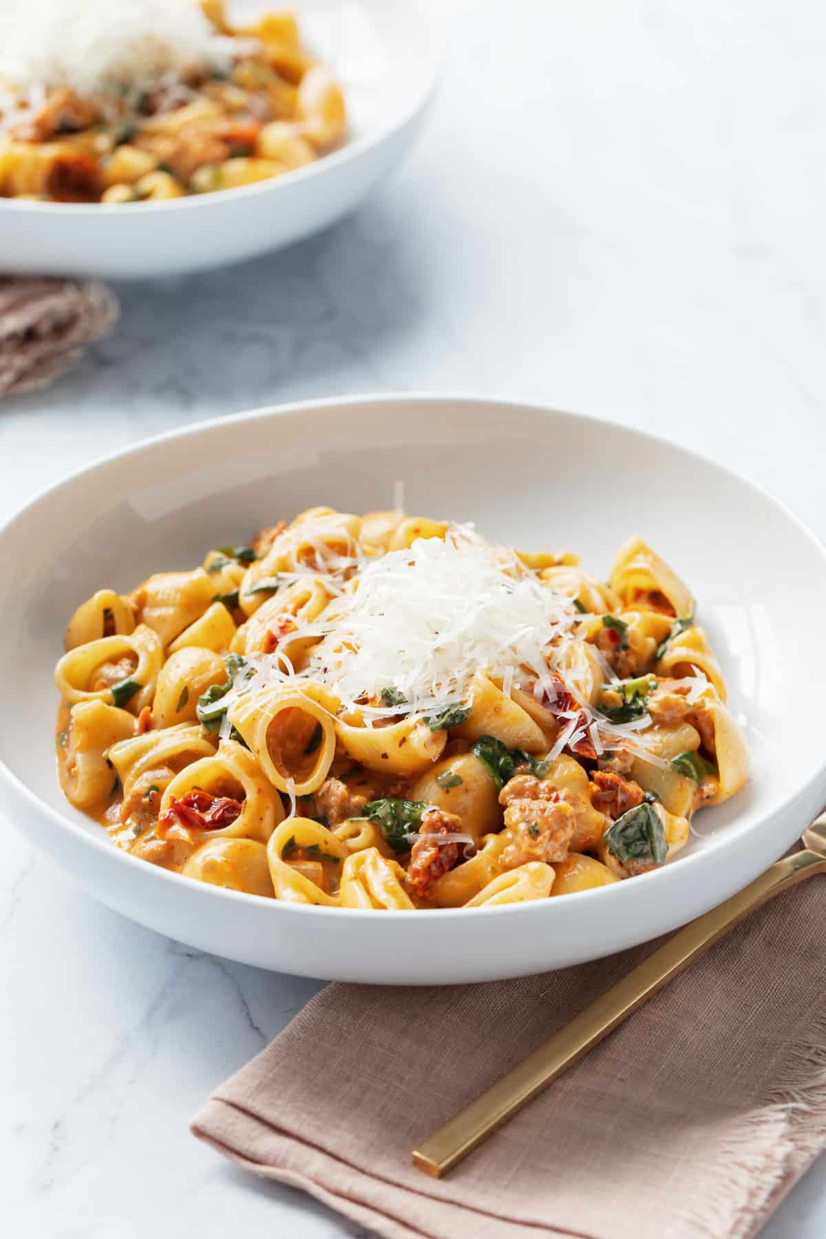 White bowl filled with Creamy Sundried Tomato and Italian Sausage Pasta and topped with finely shaved parmesan cheese.