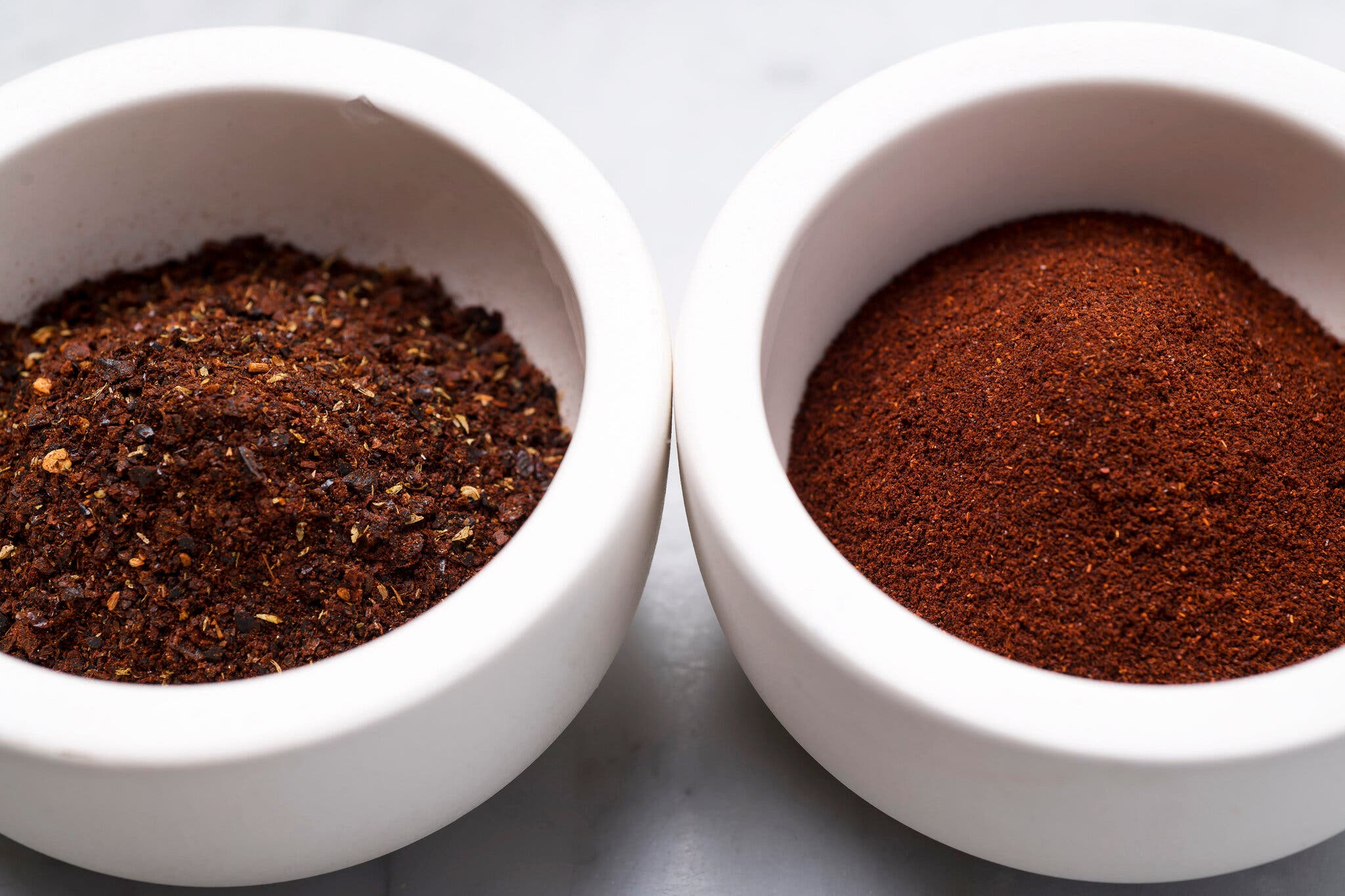 Two white bowls filled with chili powders: The version to the left is more speckled and darker than the crimson version to the right.