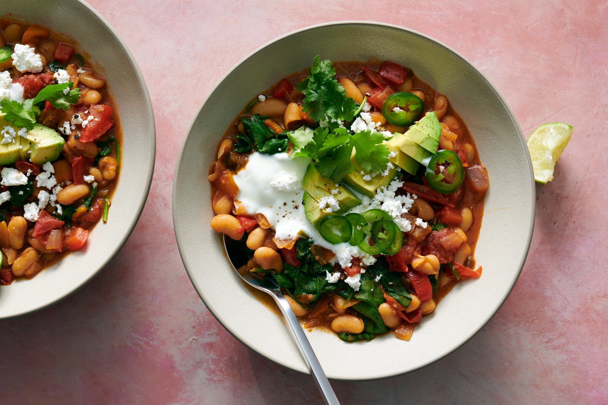 Two bowls of a white bean chili are photographed from overhead topped with sliced jalapeños, avocado and cilantro.