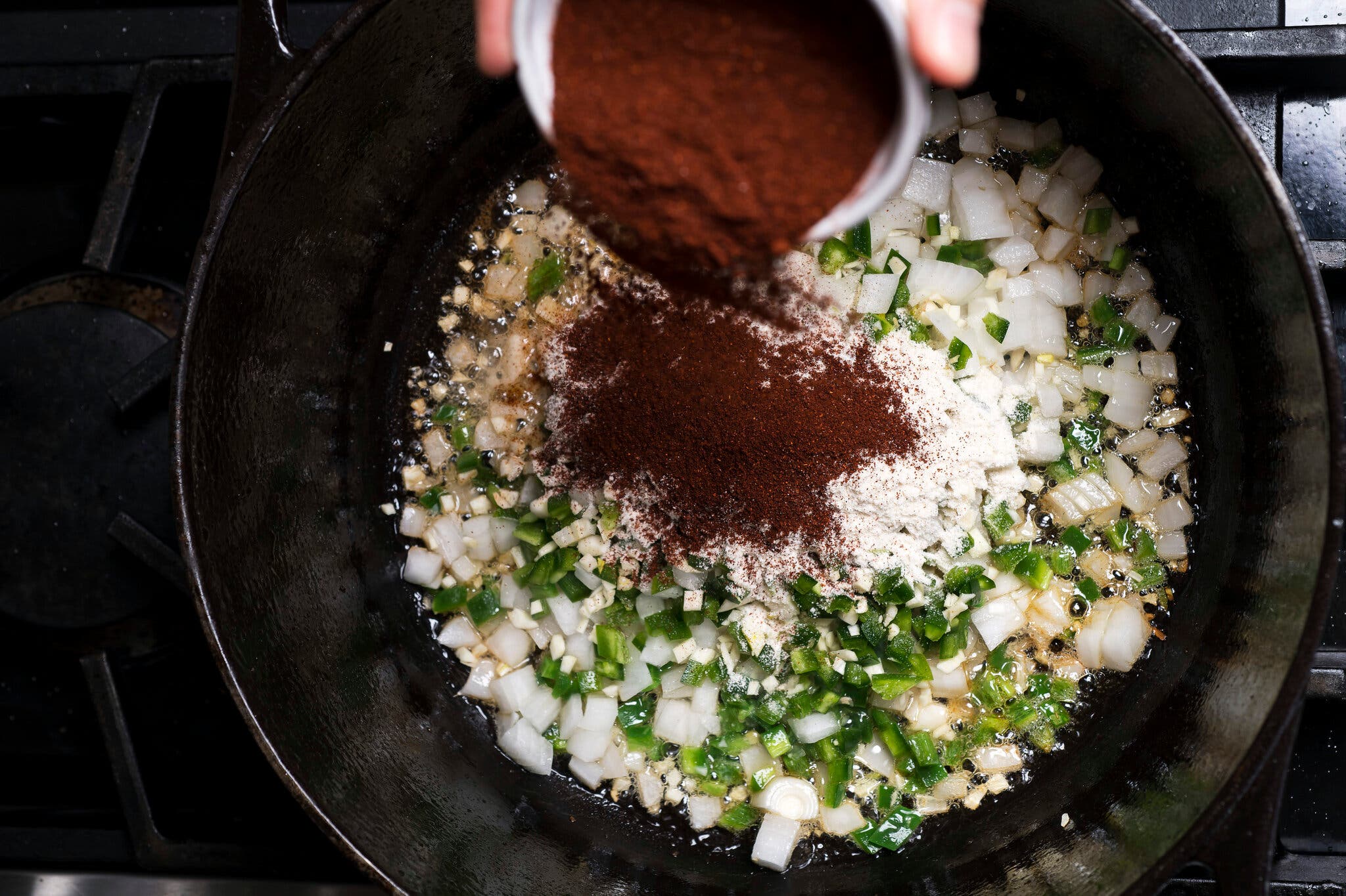 An overhead image of onions, garlic and pepper being cooked in a cast-iron skillet. A hand scatters chili powder from overhead.