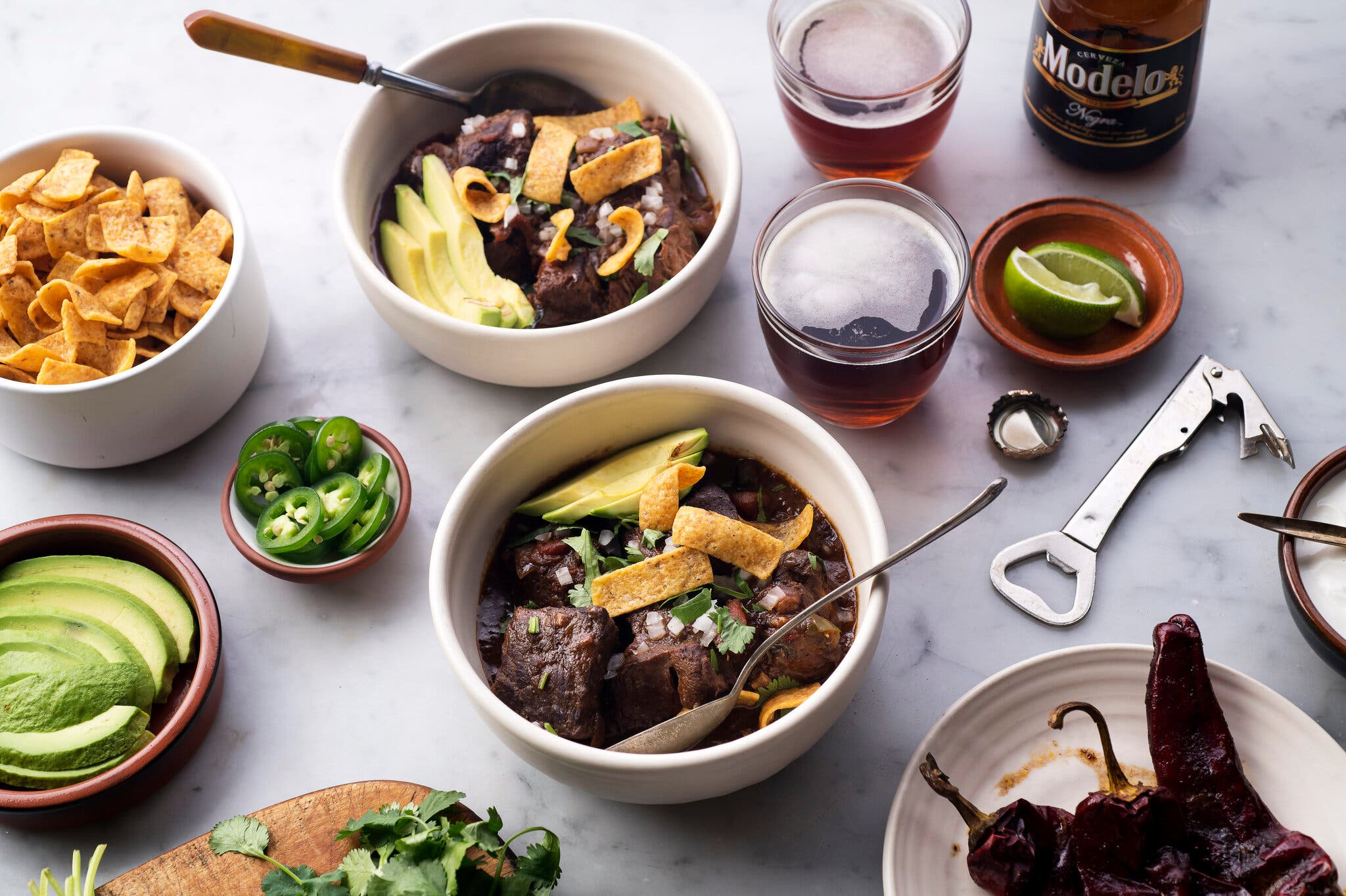 Two bowls of finished chili are topped with corn chips, avocado, onion and herbs. Bowls of the garnishes surround the servings.