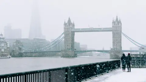 Getty Images The River Thames and Tower Bridge in London on a snowy day. In the bottom right corner there are two people dressed in dark-coloured coats and hats looking at the view behind black railings.