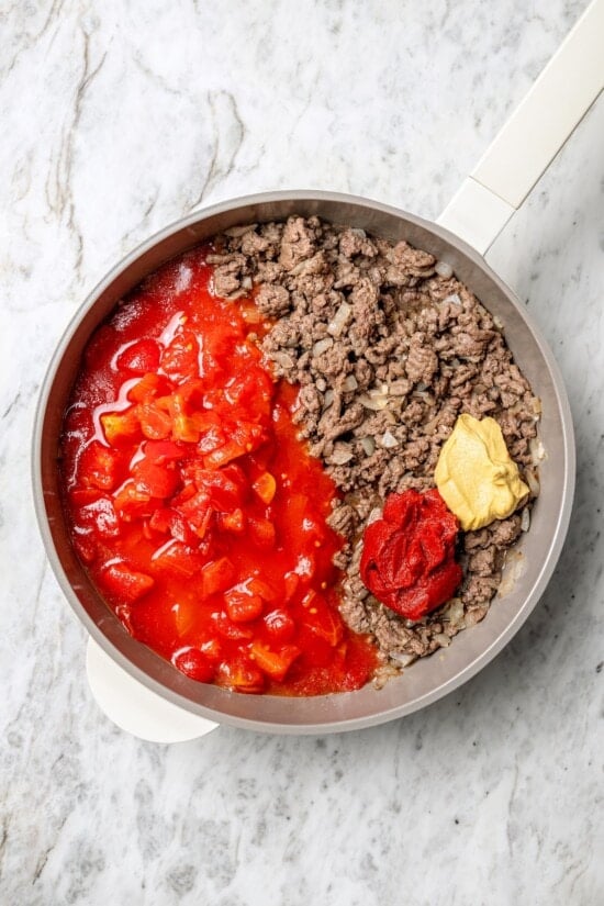 Overhead view of mustard, tomato paste, and tomatoes added to skillet of cooked ground beef