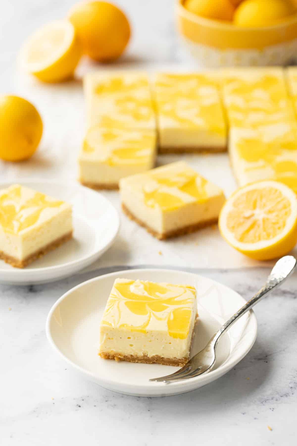 Square of Meyer Lemon Cheesecake on a small white plate with dessert fork, more cut bars and a few lemons in the background.