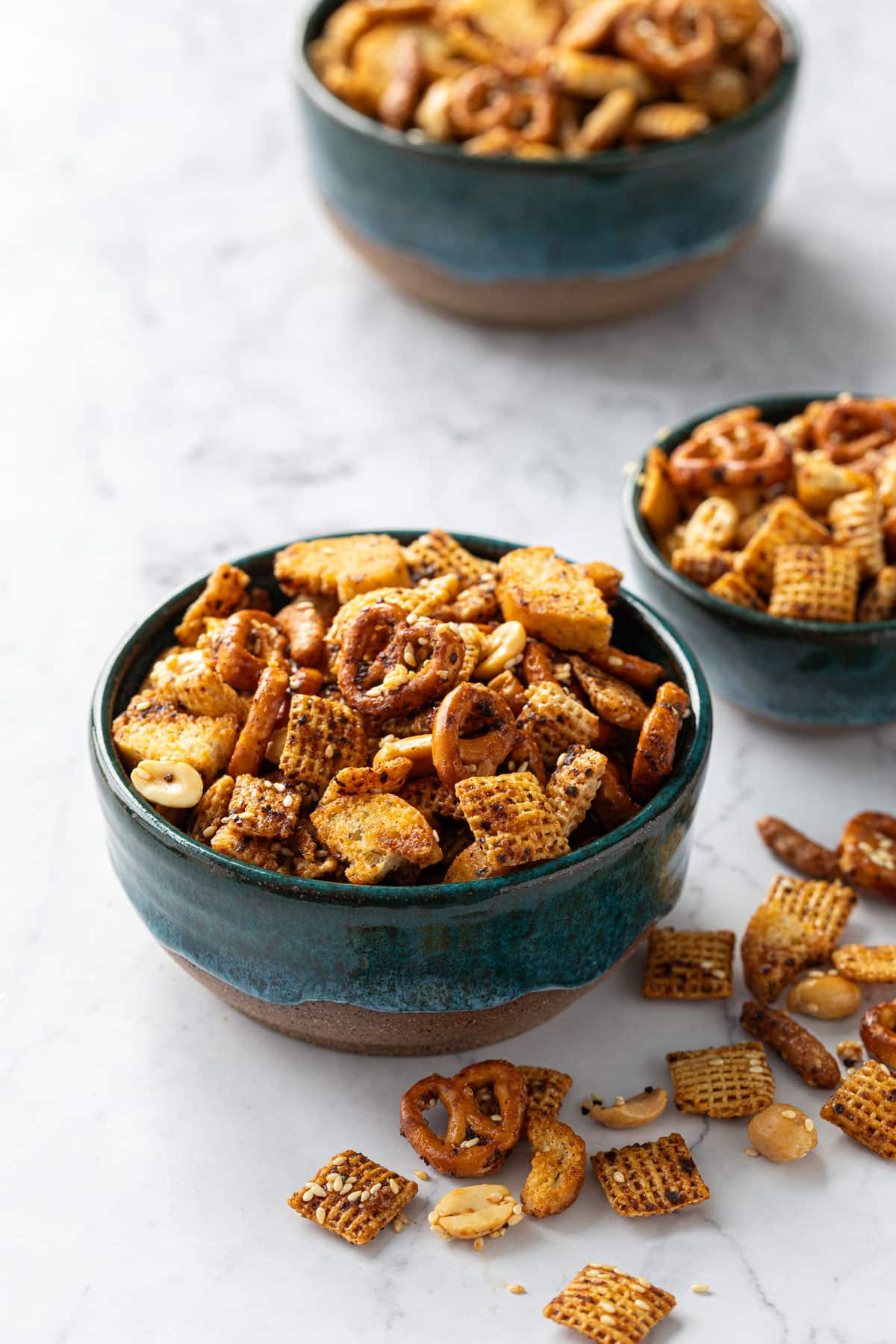 Three teal ceramic bowls overflowing with homemade Chex mix on marble background.