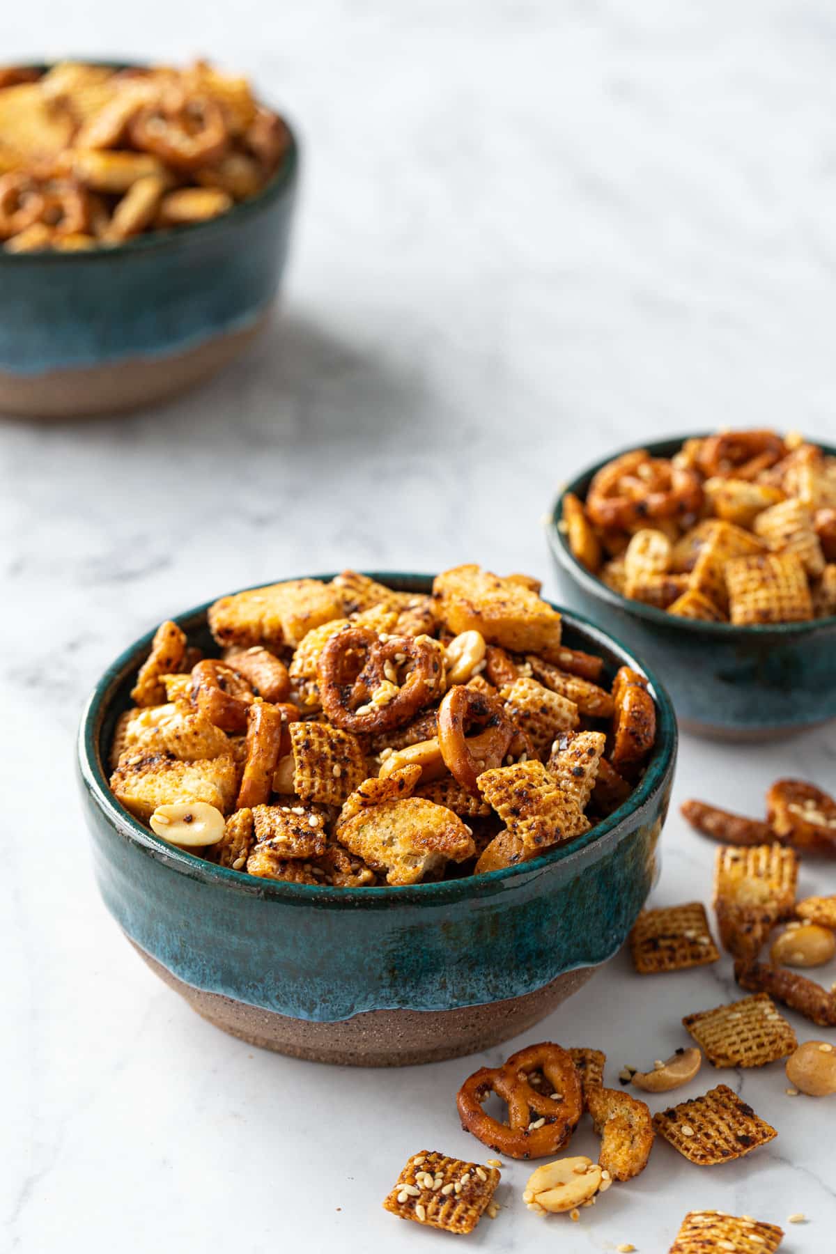 Three teal ceramic bowls overflowing with homemade snack mix on marble background.
