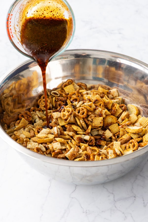 Pouring snack mix sauce mixture over dry ingredients in a large metal bowl.