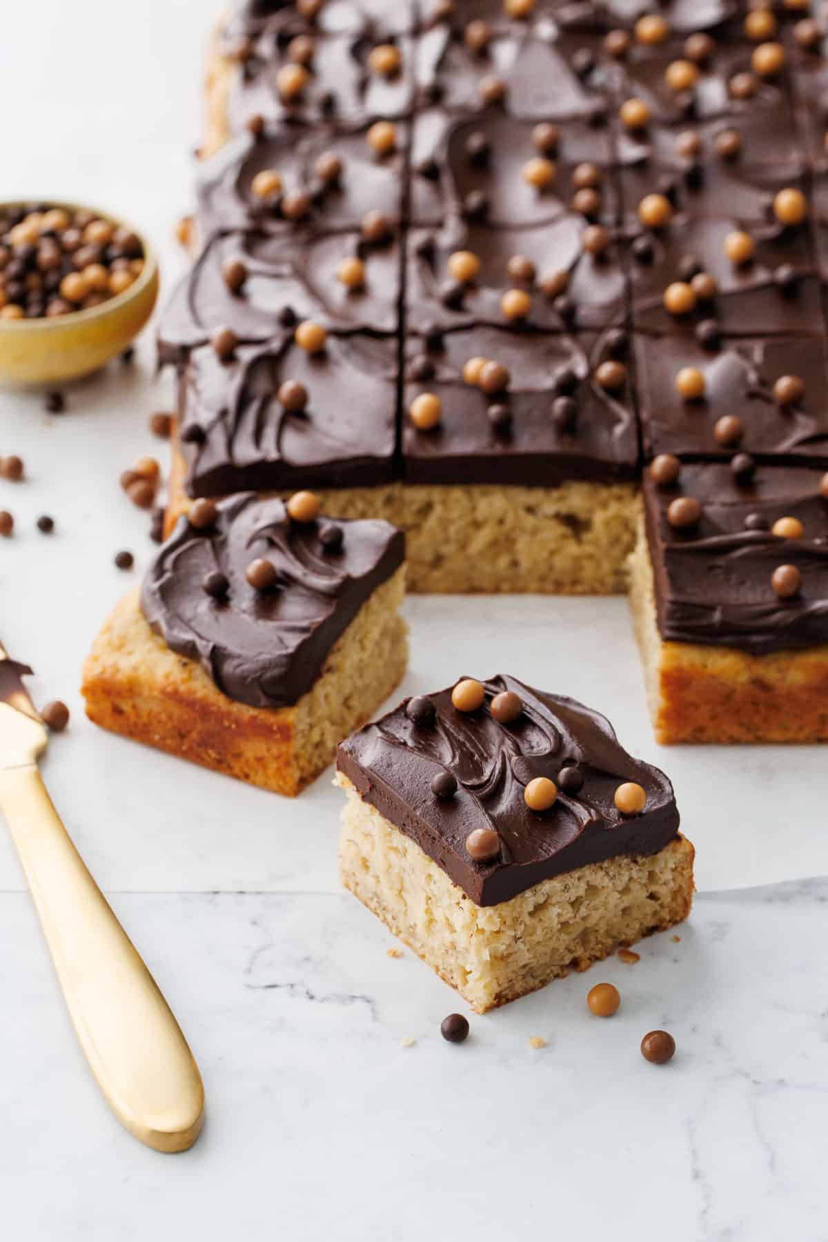 Cut pieces of Brown Butter Banana Cake with Chocolate Fudge Frosting on a marble surface with gold knife and topped with multi-color crispy pearls.