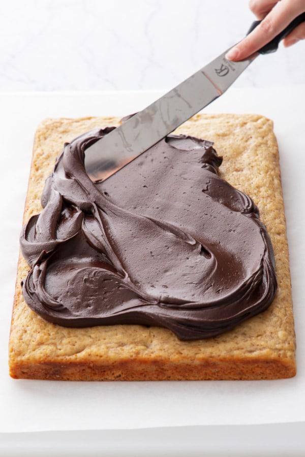 Offset spatula spreading chocolate fudge frosting on top of baked and cooled banana cake.