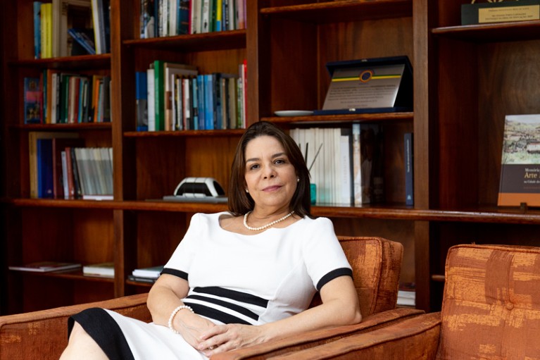 Portrait of Denise Pires de Carvalho sitting in an armchair in an office with wooden bookshelves.