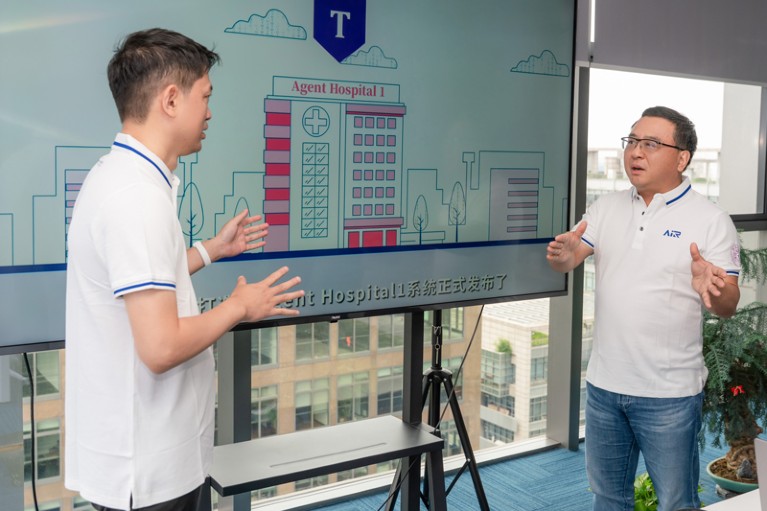 Ya-Qin Zhang and a colleague having a discussion in front of a large monitor displaying a simple line illustration of a hospital building with "Agent Hospital 1" written above it