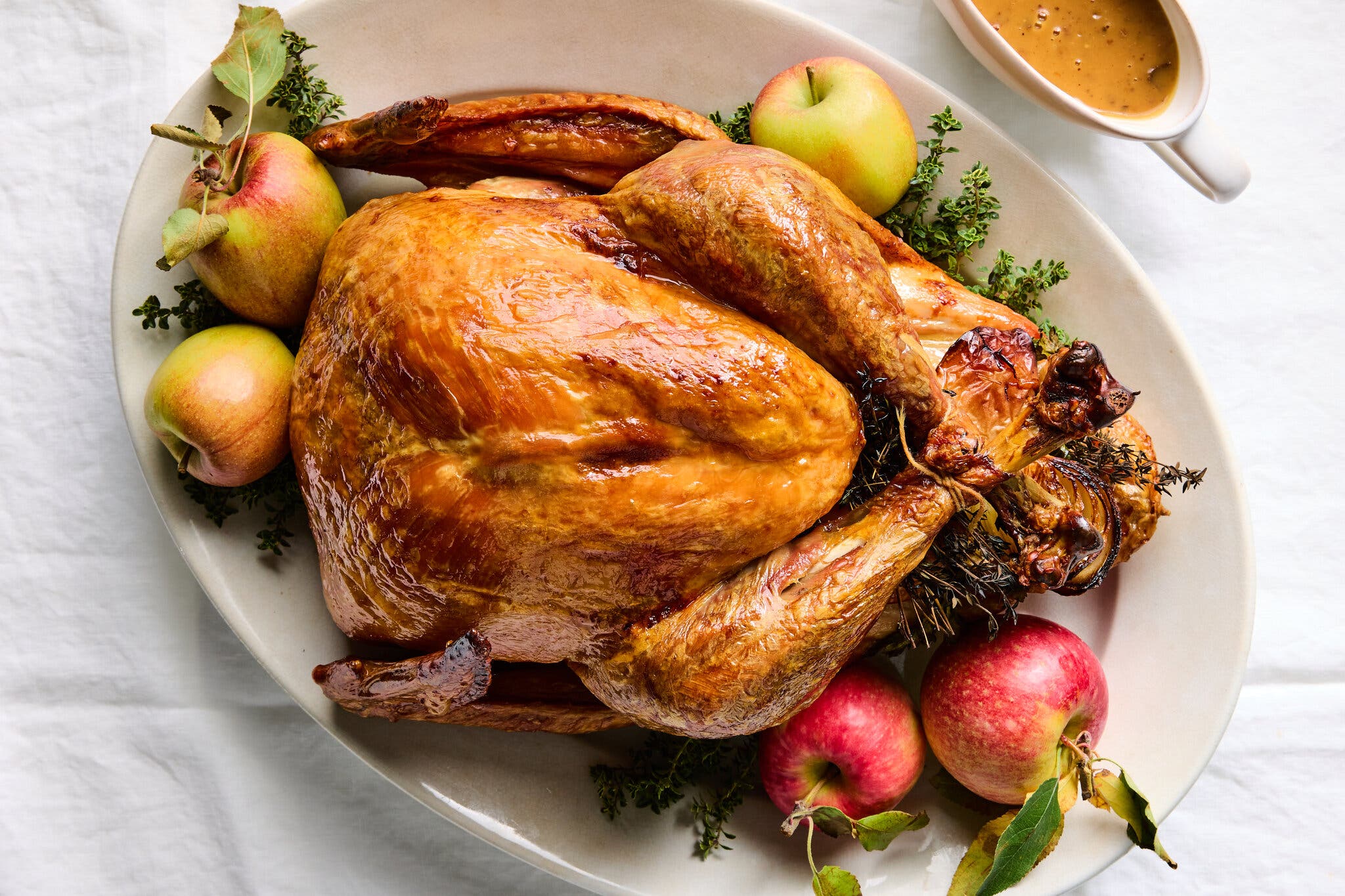 A glossy, golden-brown roasted turkey on an oval platter. Red and green apples and green herbs sit around it.
