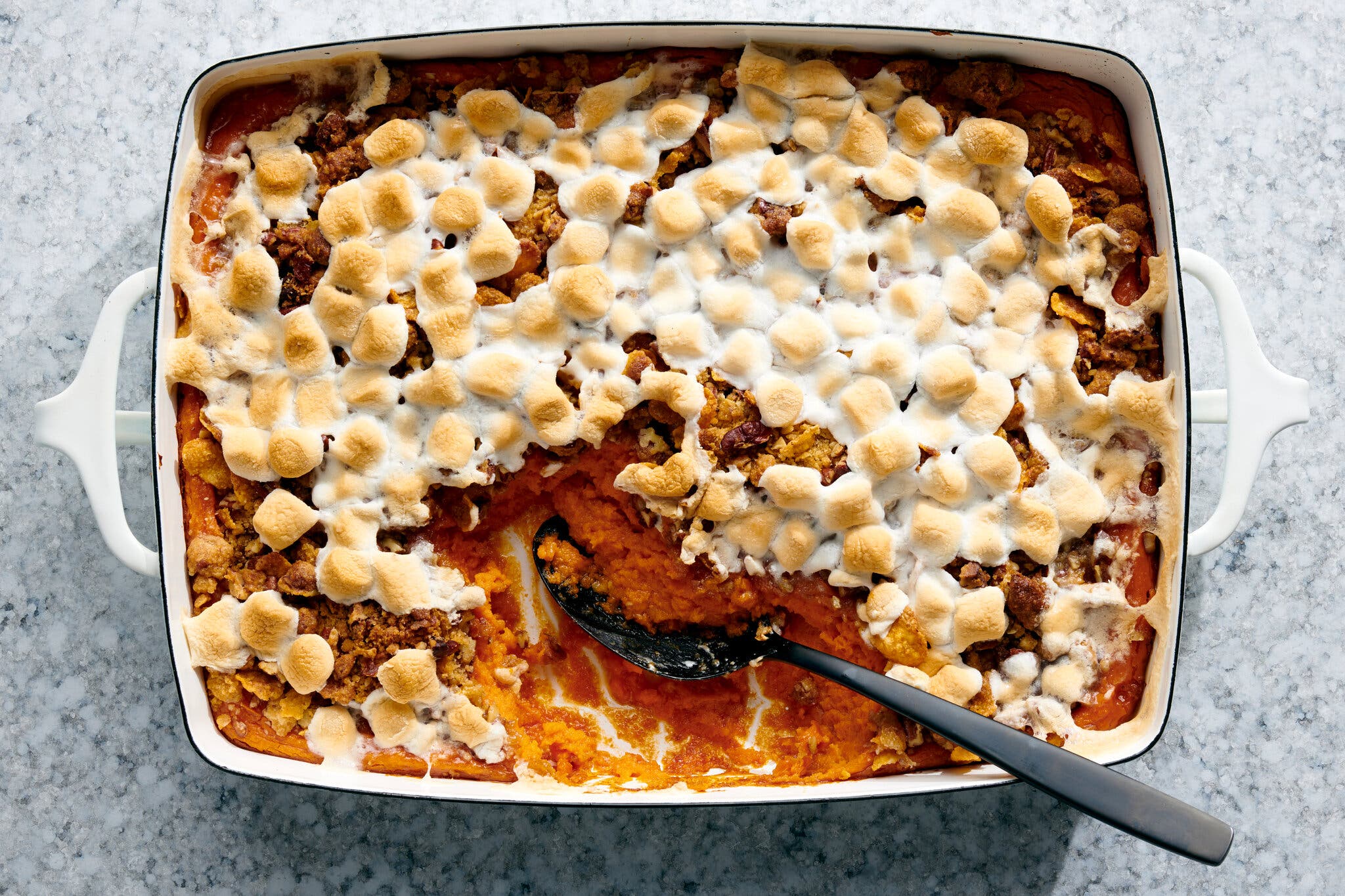 A rectangular casserole dish of sweet potatoes topped with browned marshmallows. A black serving spoon rests in a scooped-out section.