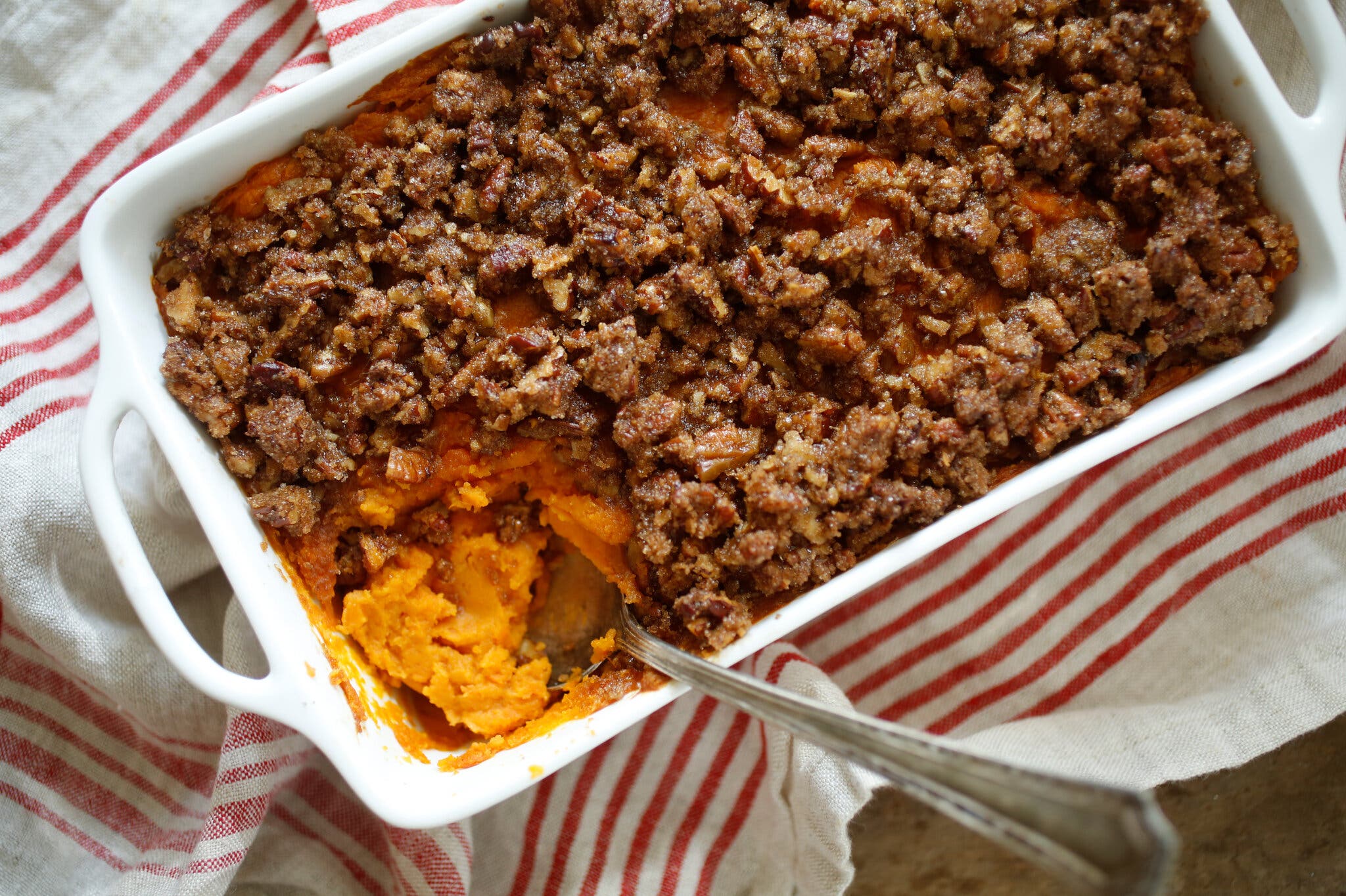 A white rectangular baking dish holds mashed orange sweet potato with a brown crumbly topping. A spoon sits in one corner.