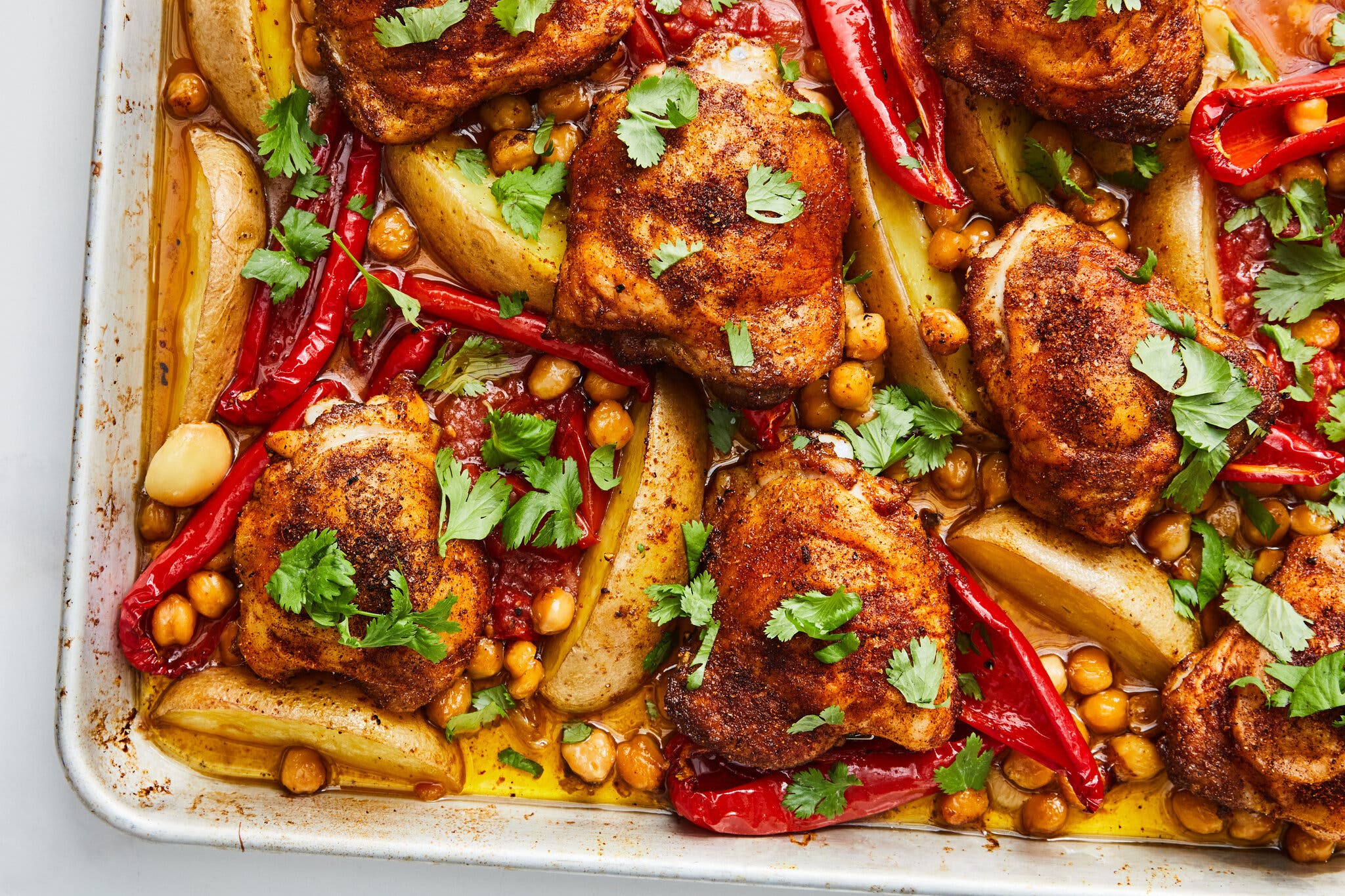 A sheet pan of baked chicken thighs, potato wedges, Romano peppers and chickpeas, garnished with fresh cilantro.