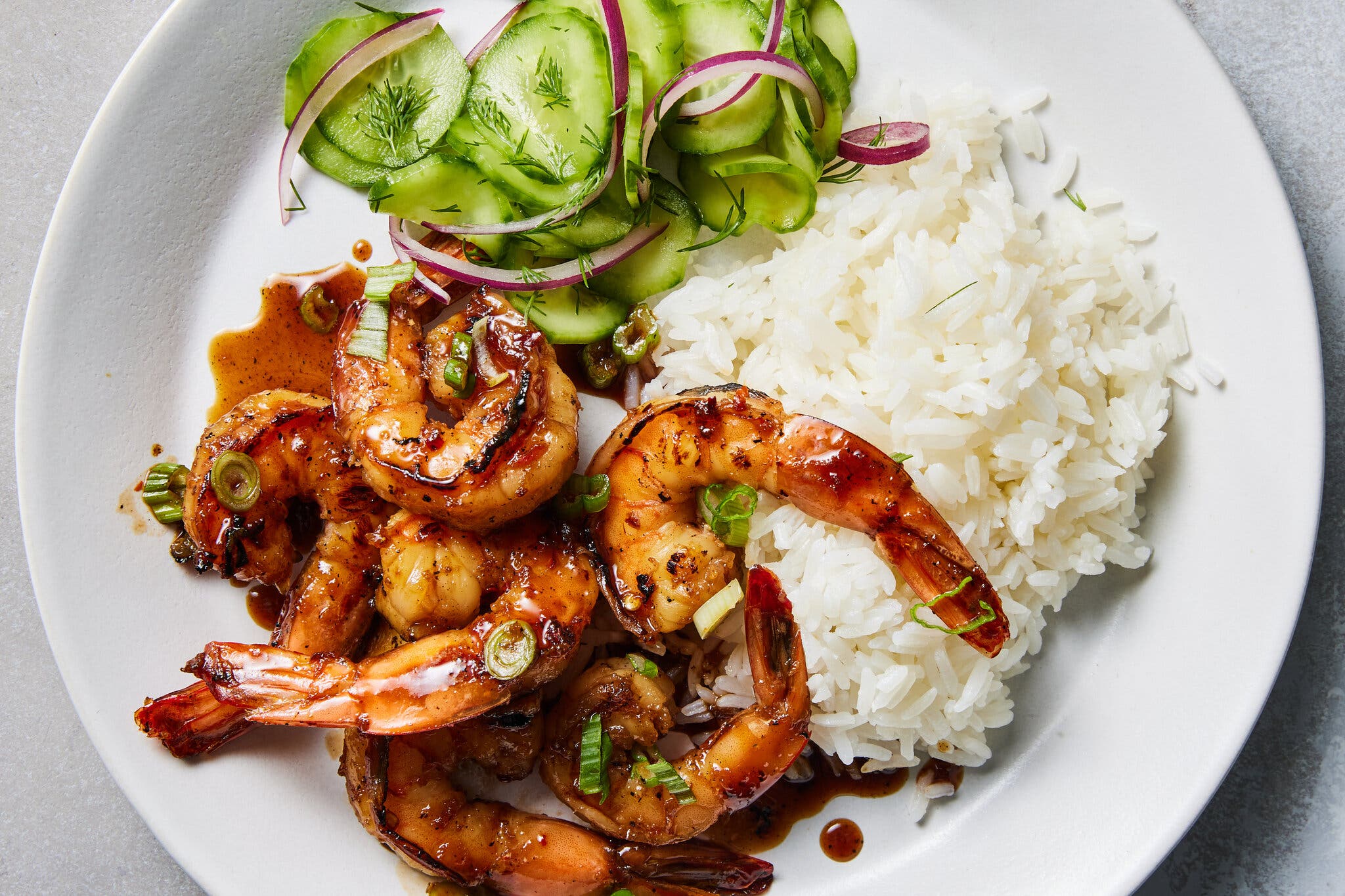A white plate filled with glazed, grilled shrimp and white rice, served with a side of sliced cucumber and red onion salad.