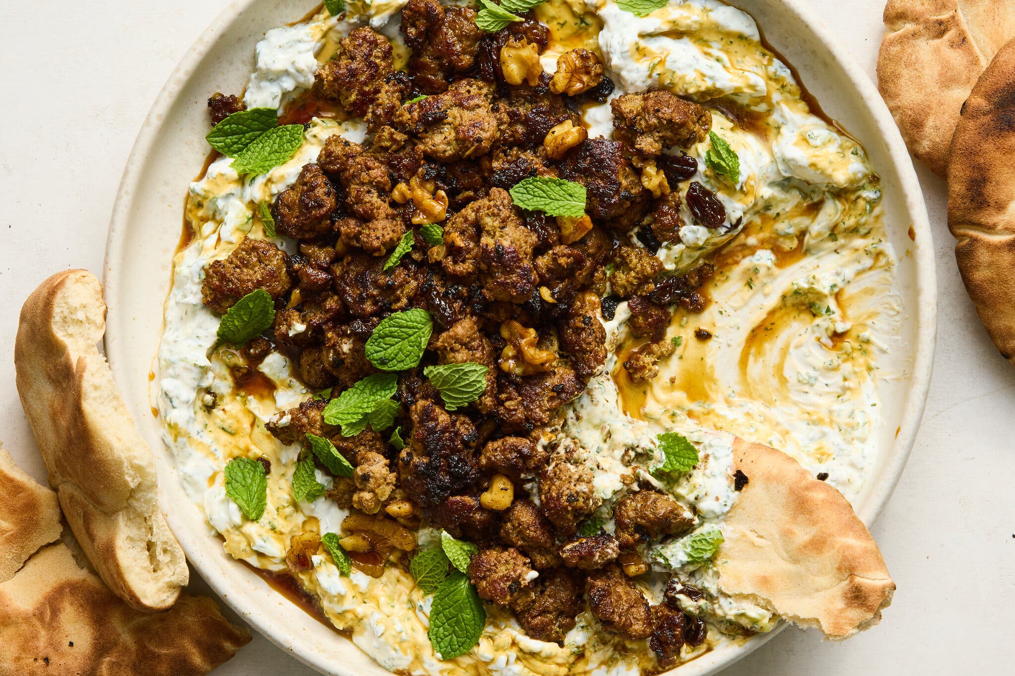 A platter of spiced ground beef, walnuts, and raisins over a bed of creamy yogurt, garnished with fresh mint, with pieces of pita bread on the side.