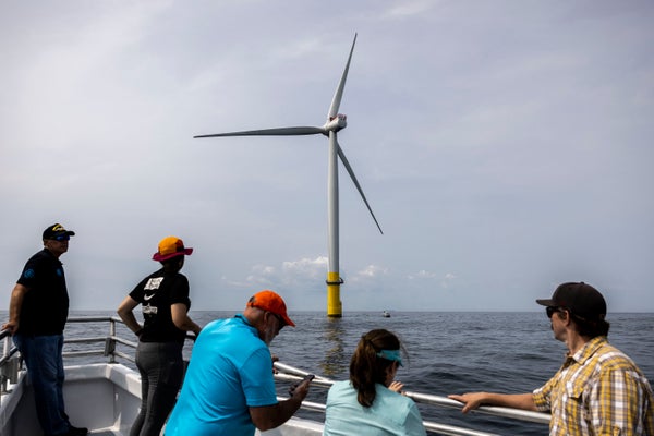 People on a boat looking at a wind turbine in the water nearby.