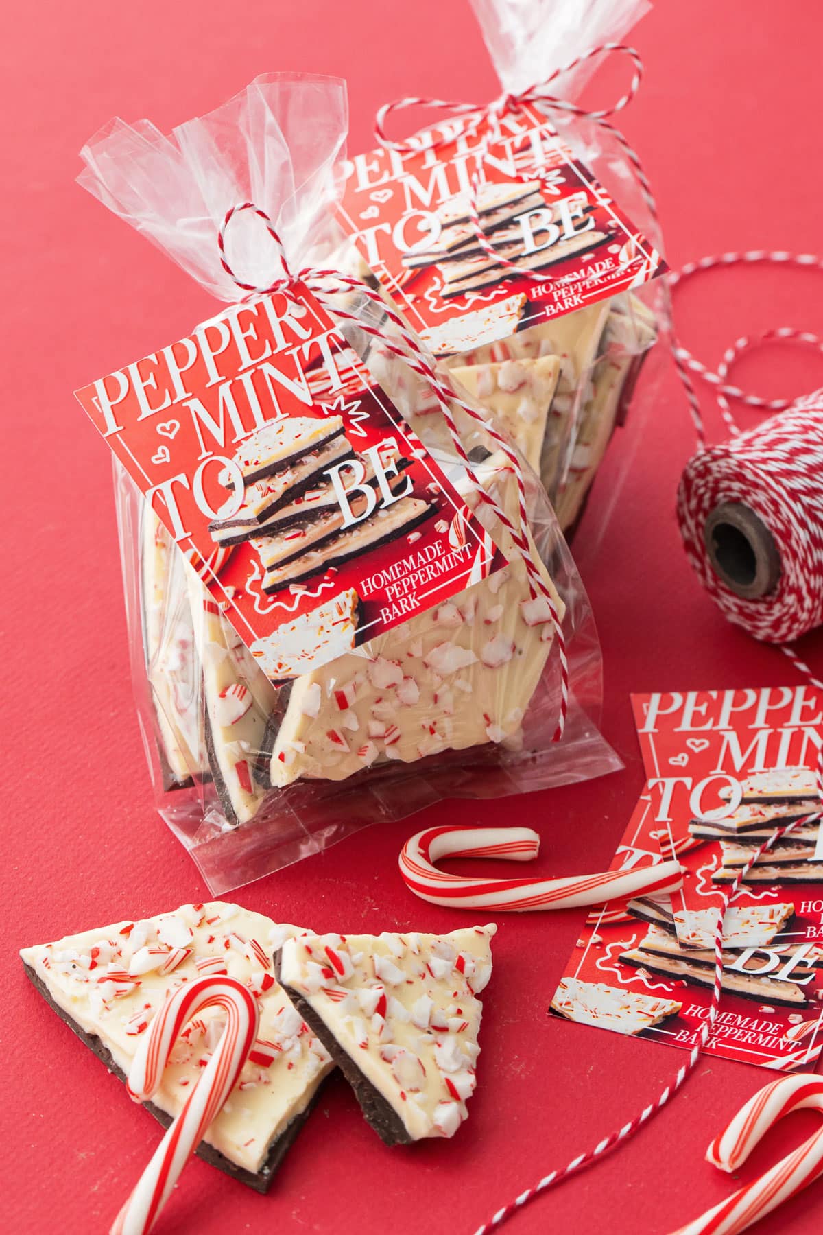 Clear treat bags filled with Homemade Peppermint Bark on a red background, tied with twine and printable gift tags.