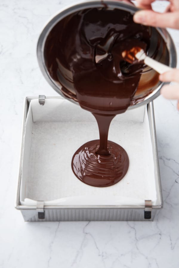 Pouring dark chocolate into a parchment-lined square baking pan.