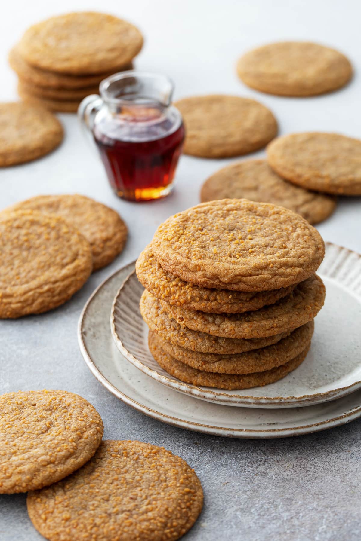 Stack of Chewy Maple Brown Butter Sugar Cookies on a ceramic plate, with a small glass pitcher of maple syrup and more cookies scattered around.