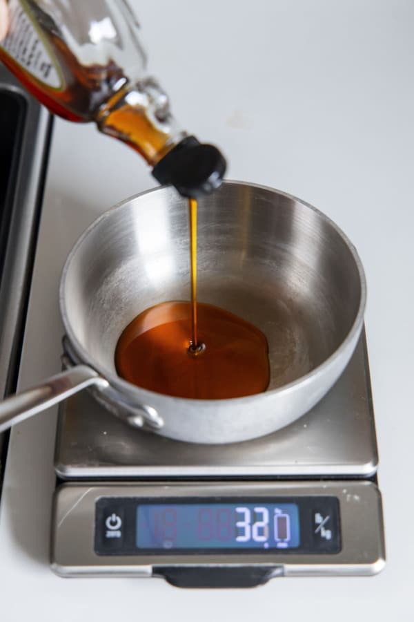 Pouring maple syrup into a saucepan set on a digital cooking scale.